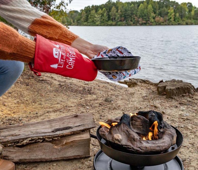 A woman leans next to a firepit with the oven mitt and pot holder. She is holding a cast iron skillet near a lake.