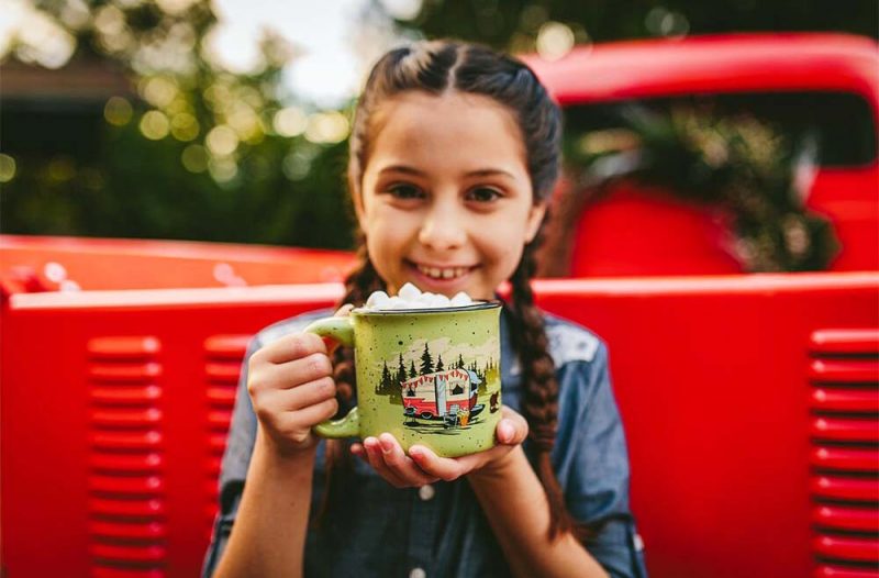 A young girl holding the Camp Casual mug. The mug is a light green with an RV and bear surrounded by trees. 