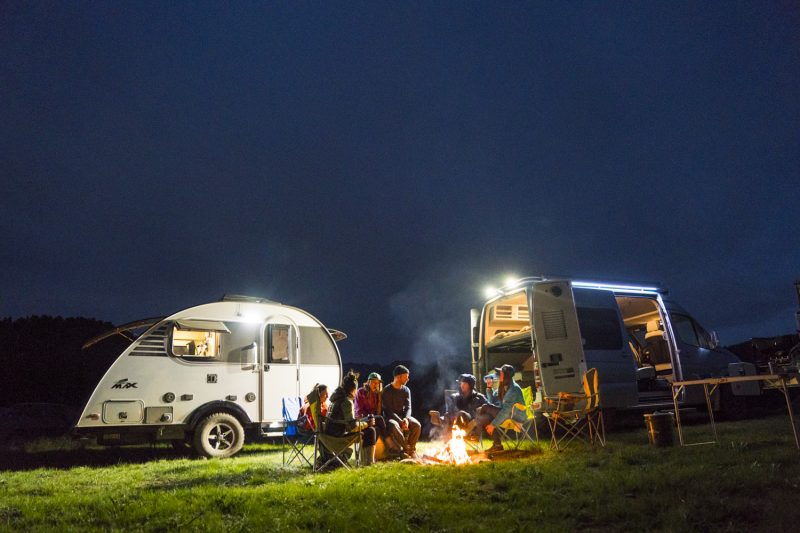 A group of people sit around the fire with a teardrop trailer and class B behind them.