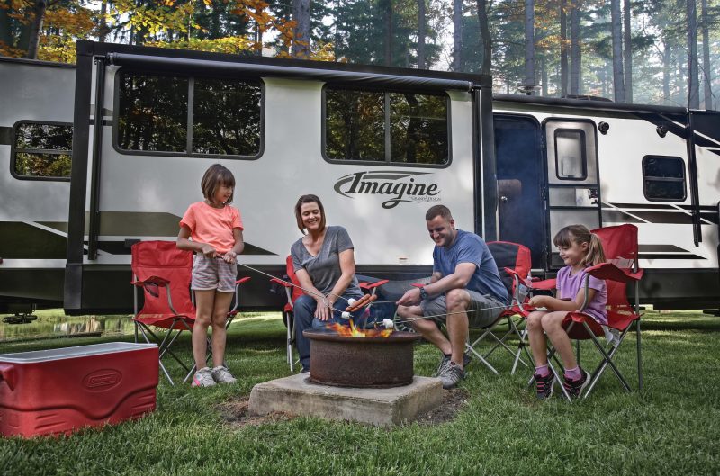 A family roasting marshmallows around a campfire with a travel trailer behind them.