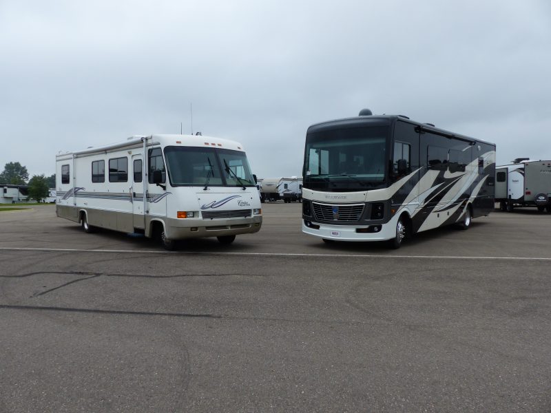 A used motorhome is parked to the left of a new motorhome.