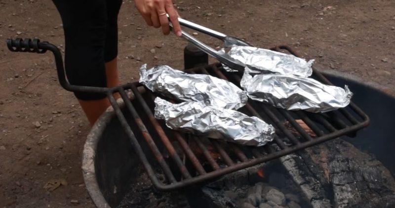 A woman using tongs to turn the Cheesy Camping Hot Dogs over the campfire.