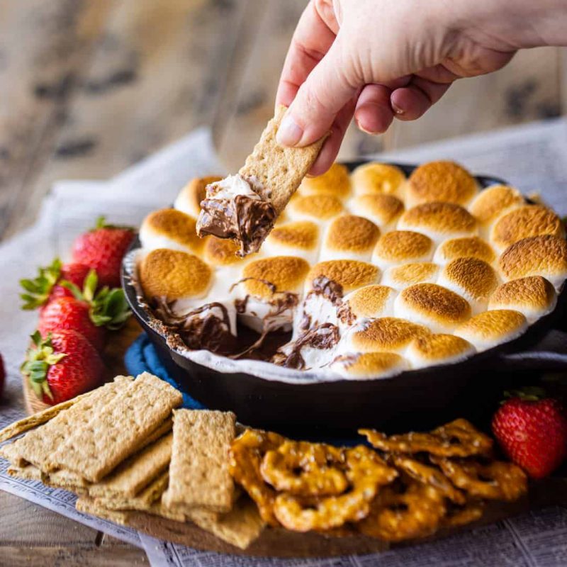Someone dipping a graham cracker into the finished Campfire S'mores Dip. A variety of crackers and strawberries are laid around the skillet.
