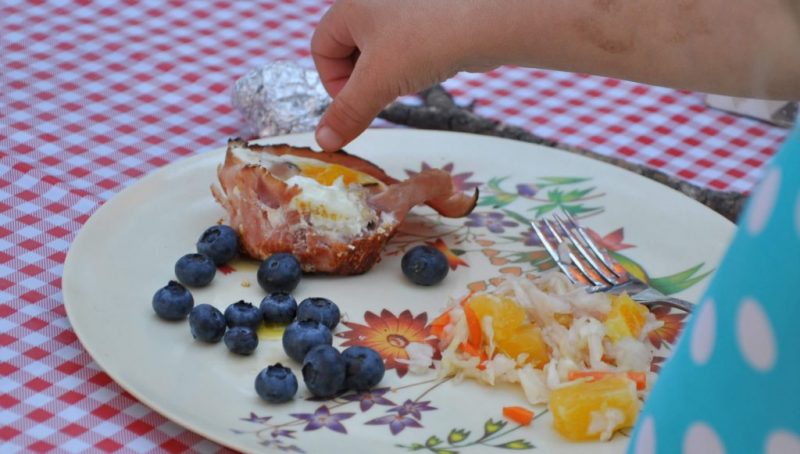 A child enjoying an Egg Cup with Ham with blueberries and veggies on a plate.