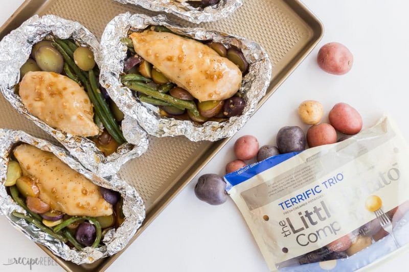 Four Honey Garlic Chicken Foil Packets being prepped on a baking tray with a bag of potatoes near it.