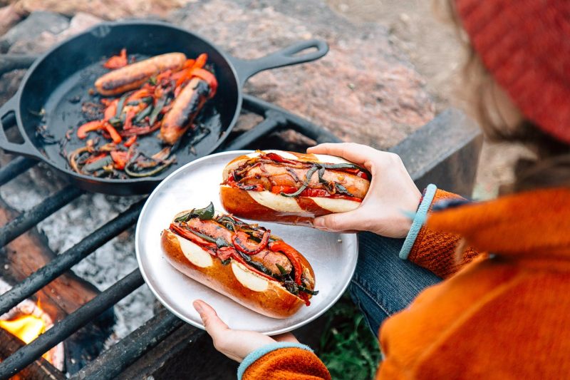 A woman enjoying Brats with Peppers and Onions near a campfire.