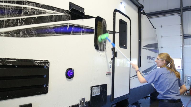 A woman cleans an RV's exterior with a long-handled soft bristle brush.