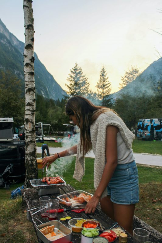Woman serving food in banquet-style tins with steam rising rapidly, a number of containers and bowls of various foods across the table in front of her. Campsites in the background.