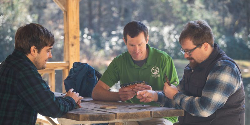 Three men sitting at a picnic table playing 10 Essentials card game.