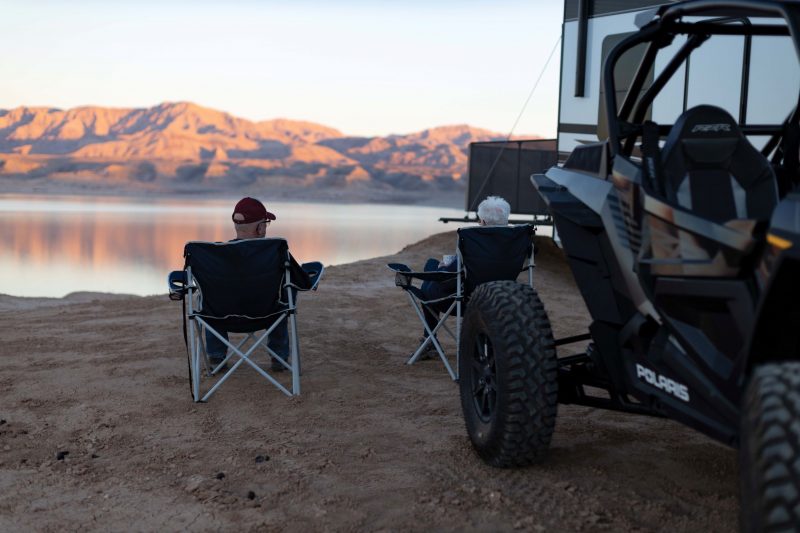 A couple is sitting in camping chairs near their Momentum toy hauler and ATV. They are watching the sunset near a lake with mountains in the distance.