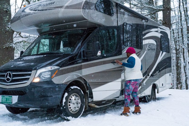 A woman wearing a winter cap, vest, colorful leggings and waterproof boots opens the door of her Class C motorhome in a snowy, wooded area.