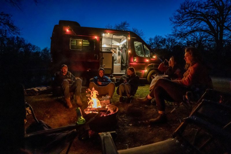 A group of friends are sitting around the campfire, the motorhome providing light in the background.