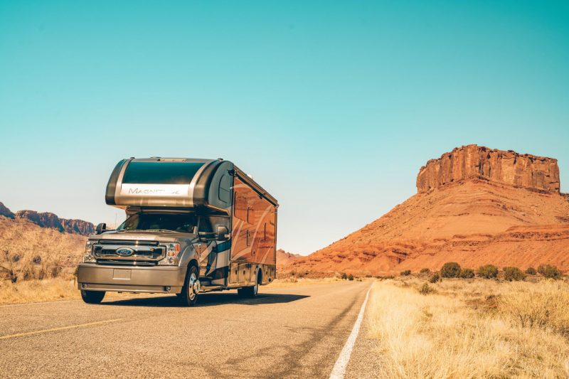 A Class C motorhome travels along a desolate road in a desert landscape of the western United States.