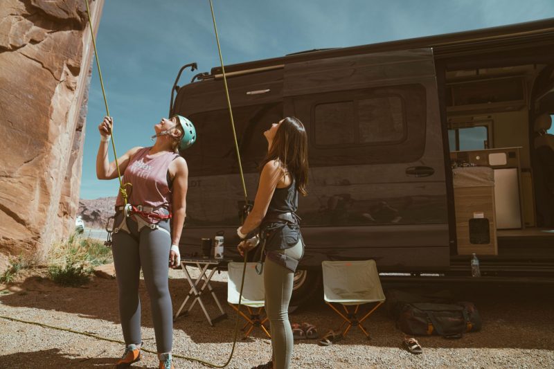 A still scene from one of Becky's B Van Adventures videos shows two women wearing rock climbing gear as they stand before a rock wall in a western United States desert. Behind them is a camper van acting as their mobile basecamp, complete with outdoor chairs and a table.