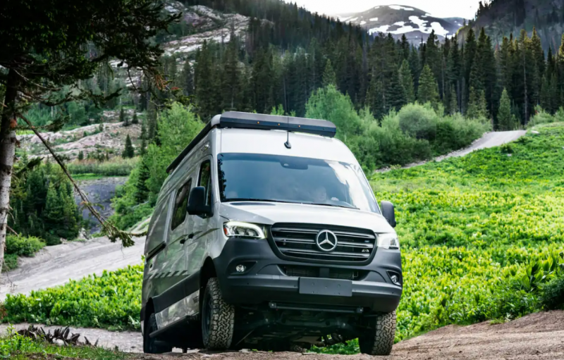 A Winnebago Revel camper van climbs steep terrain. Lush green vegetation, evergreen trees, and a snow capped mountain are visible behind the RV.