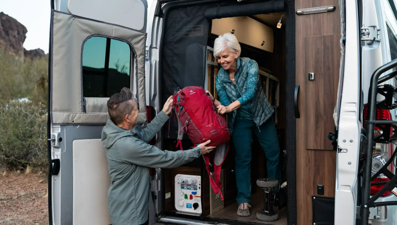 A woman hands gear to her partner as they unload their camper van.