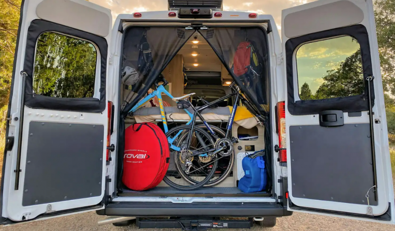 The rear doors of a Winnebago Solis Class B RV are opened and show the RV packed with two bikes and additional outdoor gear.