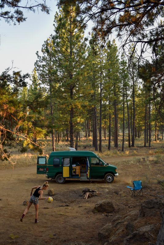 A woman plays with a ball near a dark green camper van parked in a wooded forest campsite.