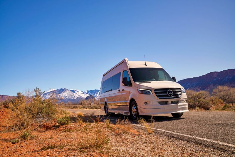 A white Class B motorhome travels along a highway surrounded by desert vegetation and distant …