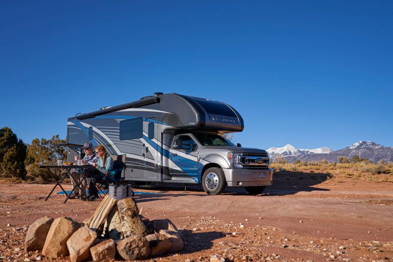 A Class C motorhome in the desert. The RV is parked on reddish brown dirt. In the far background, snow-capped mountains rise towards a clear blue sky.