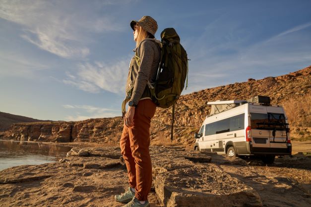 A woman wearing an outdoor pack and hiking apparel stands near a Thor Sequence camper van.