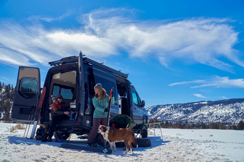 A man, woman and dog prepare for snowy outdoor activities. The man is sitting inside the rear of the Class B RV as he puts on his shoes while the woman holds onto skis. 
