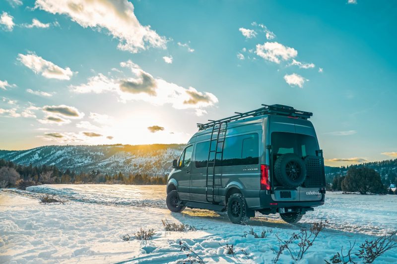 A Tiffin Cahaba Class B RV goes off road in snowy terrain in a scene captured during Becky's B Van Adventures. In the distance, mountain slopes are covered in snow and evergreen trees.