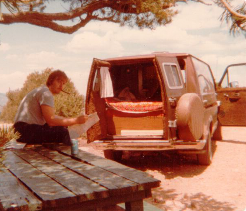 An aged photo of a man with a cigar sitting on a picnic bench with a camper van behind him. The camper van's doors are open revealing a bed in the back and curtains over the windows.